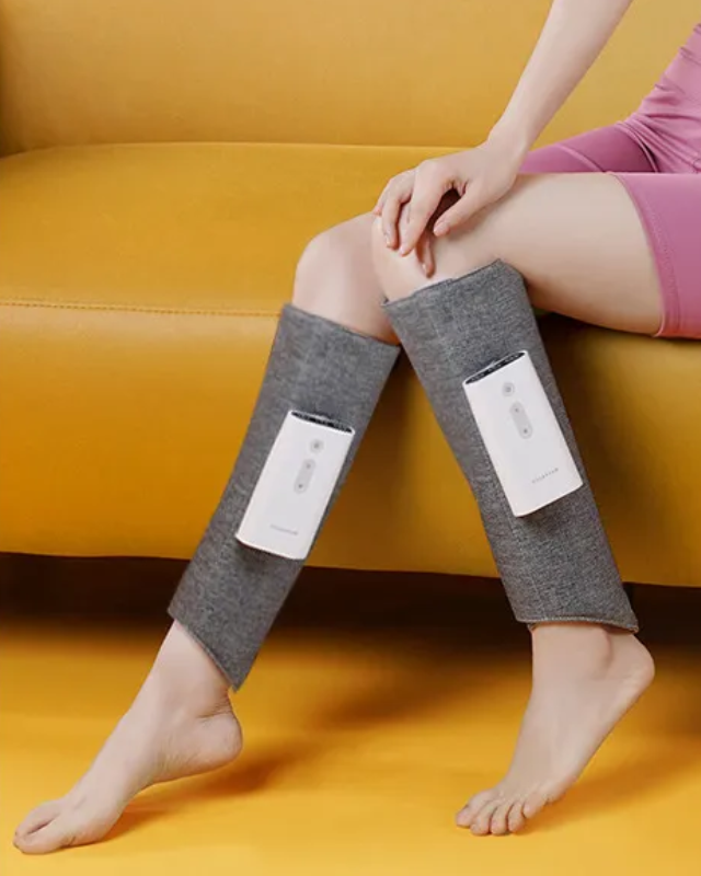 Woman wearing a gray heated leg massager on both legs while sitting on a yellow sofa